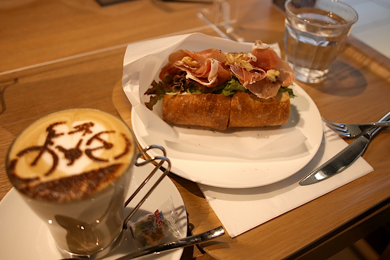 フランスパンに生ハム、クリームチーズ、レタスが挟まれたサンドイッチと、自転車のラテアートが施されたカフェラテ